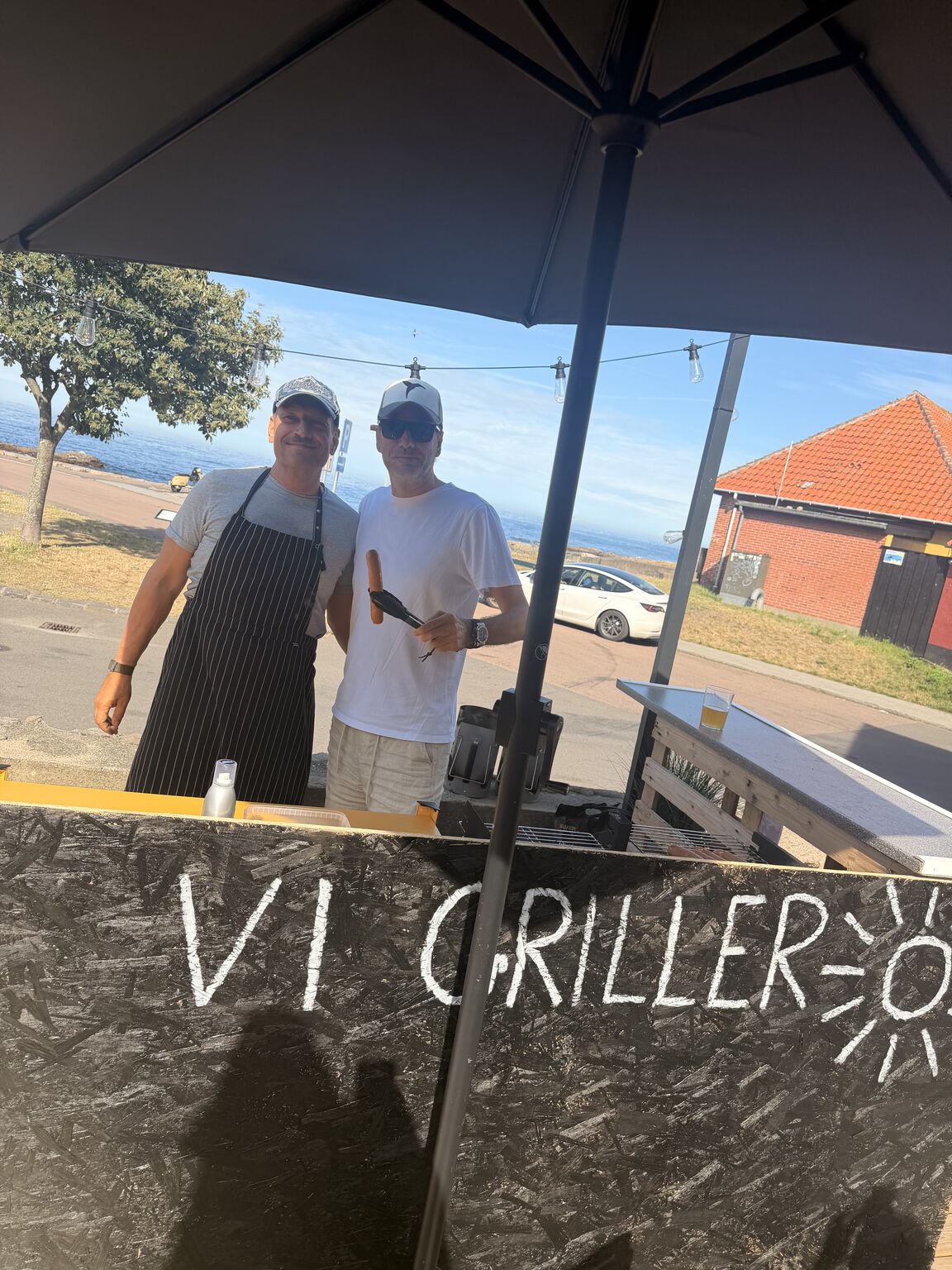 Martin Thorborg og Jesper Buch står bag en grillbod på havnen i Gudhjem en solrig sommerdag. Martin har stribet forklæde og kasket på, Jesper har hvid t-shirt, hvid kasket og solbriller på og holder en pølse i grilltangen.